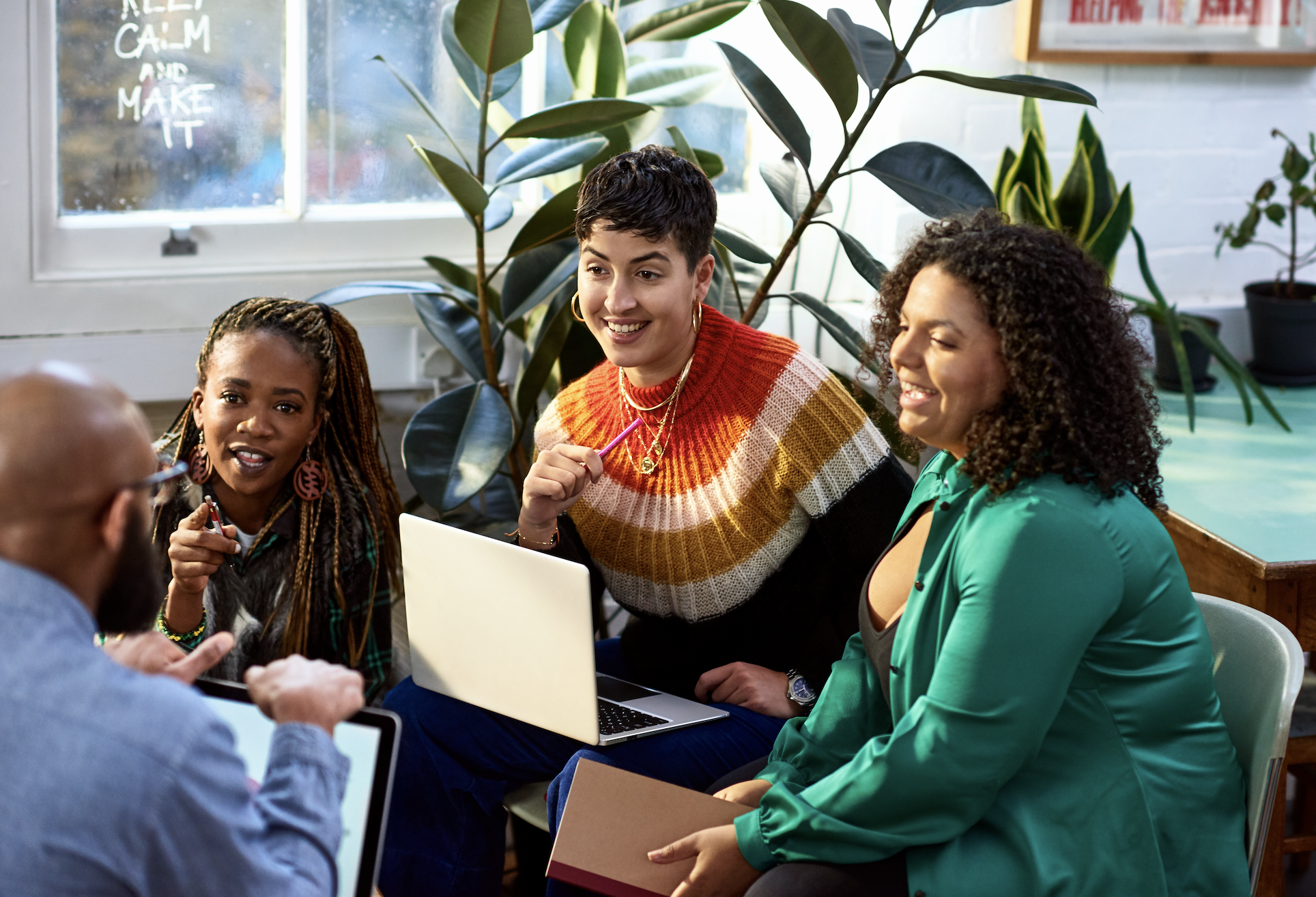 Four people engaged in a discussion are seated in a room with plants, laptops, and books around them. They appear to be happy and focused