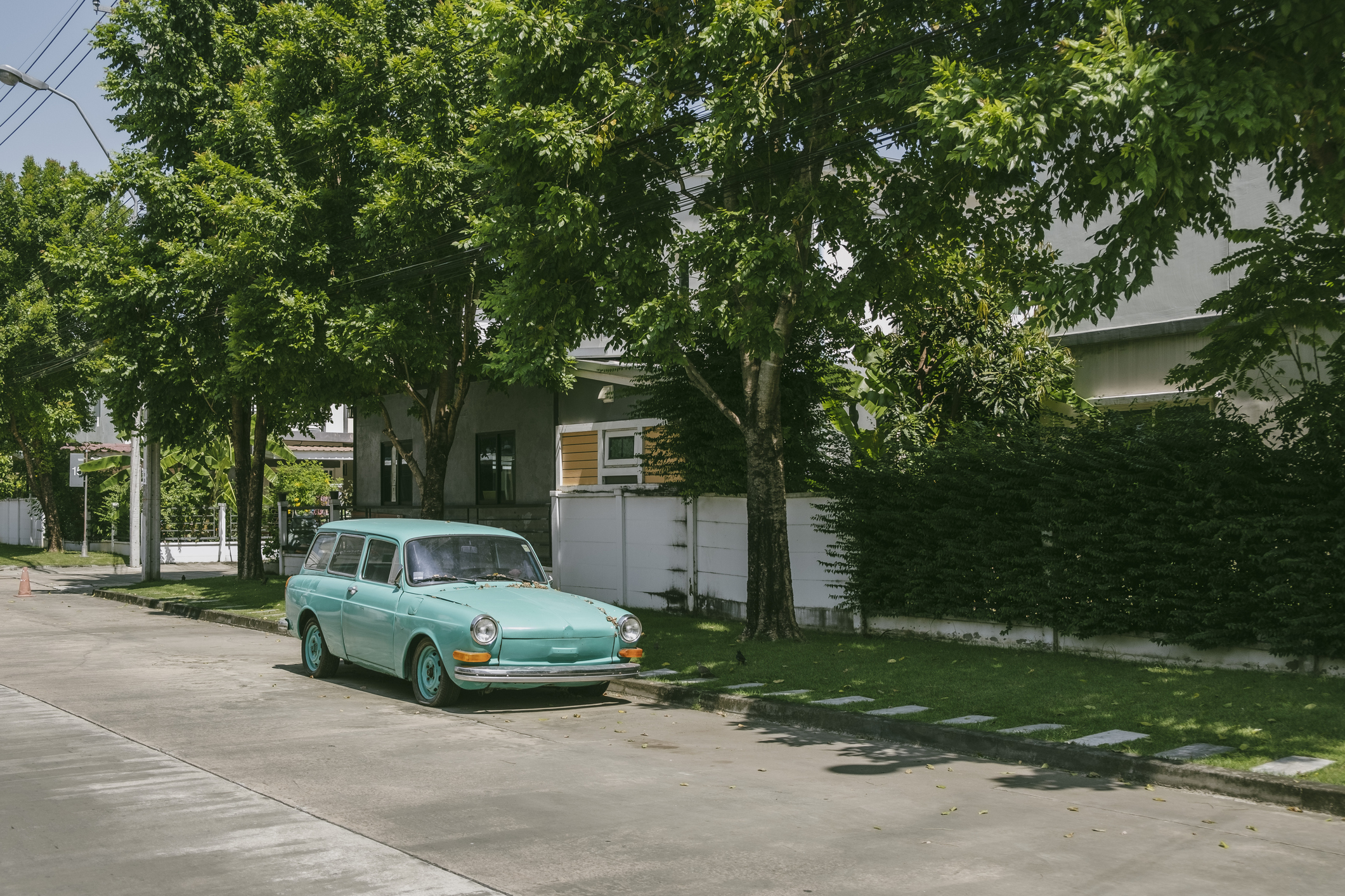 A teal vintage car is parked on a residential street with trees lining the sidewalk and houses in the background