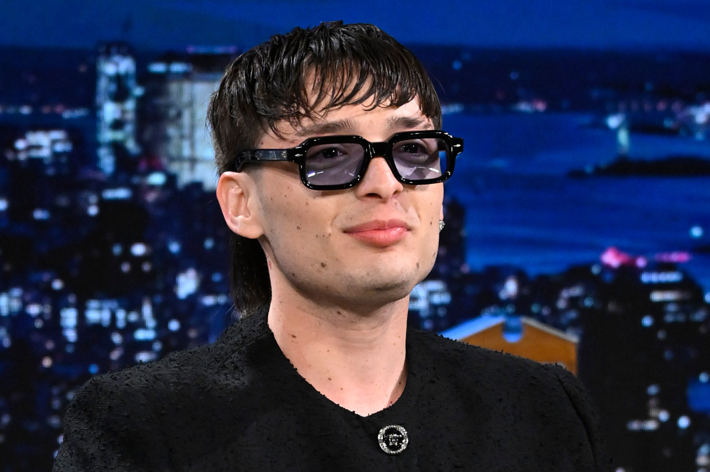 Bad Bunny wearing black, textured jacket with a brooch and dark sunglasses, nighttime cityscape in the background