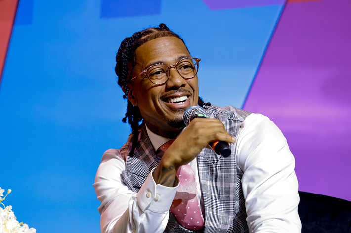 Nick Cannon speaks into a microphone at a music event, wearing a plaid vest, white shirt, and pink tie, with a flower arrangement beside him