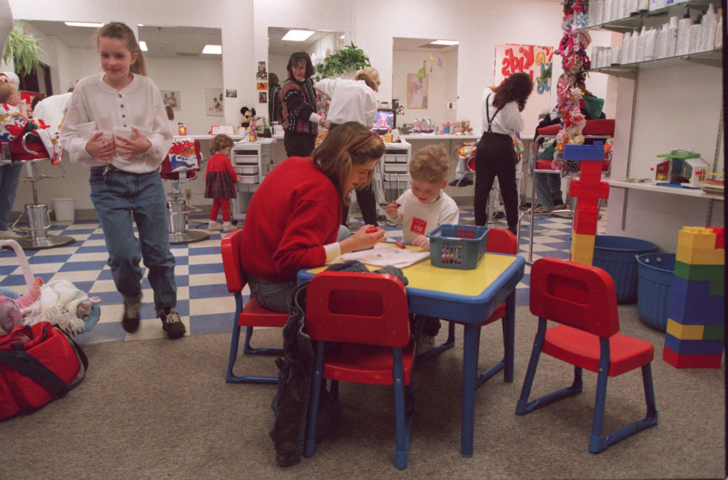 A classroom with children and adults engaged in activities. A woman helps a child with schoolwork at a small table, and another child walks in the background