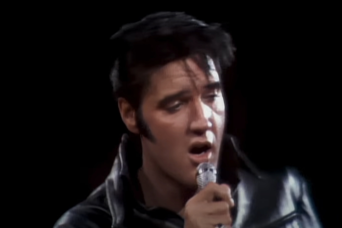 Elvis Presley sings into a microphone, wearing a black leather jacket