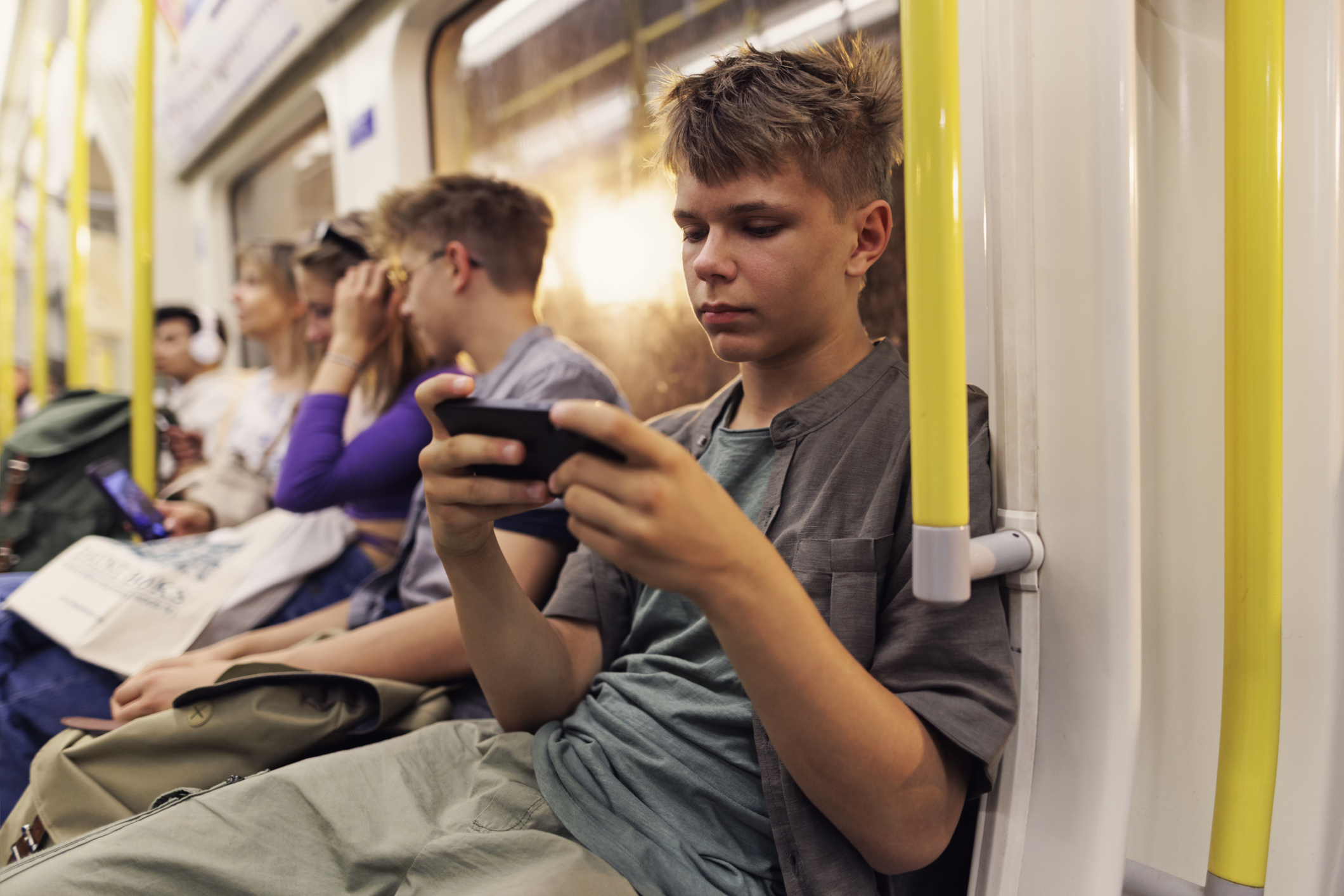 A teenage boy sits on a subway, engrossed in his phone. Other passengers are also absorbed in their devices, creating a sense of individual focus in a shared space