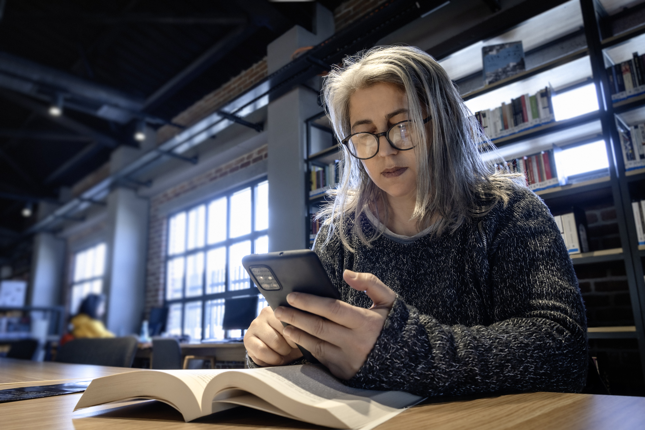 Woman with long hair, glasses, and a sweater reading on her phone in a library, an open book on the table. No other persons named