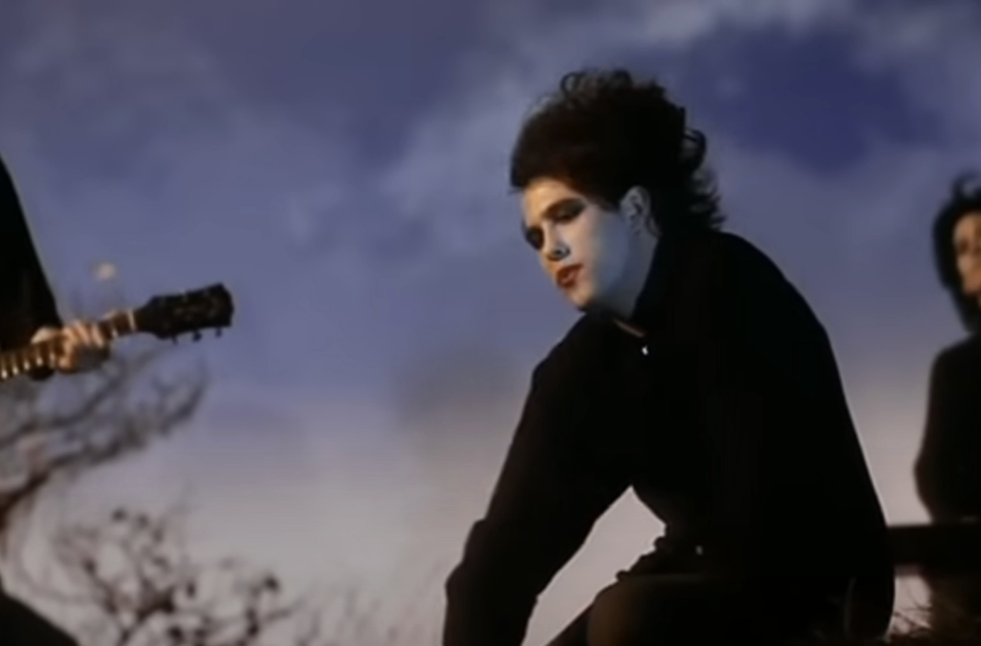 Robert Smith from The Cure in a music video, dressed in signature gothic style with heavy makeup and dark clothing, performing outdoors with fellow band members