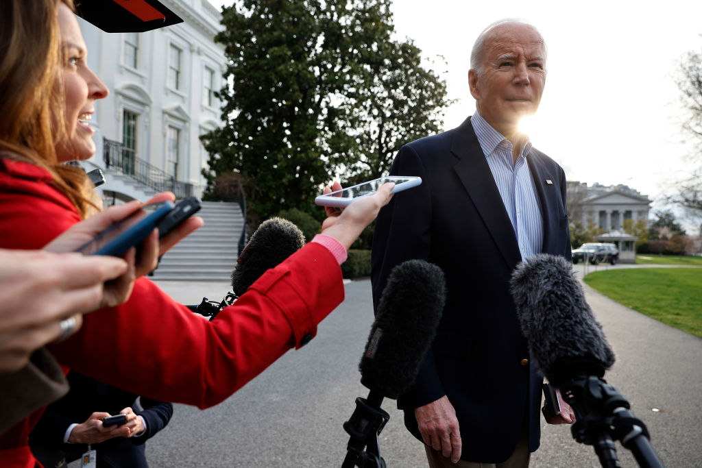 Journalists surround Joe Biden outside the White House, holding microphones and recording devices near him as he speaks. Biden wears a dark suit jacket