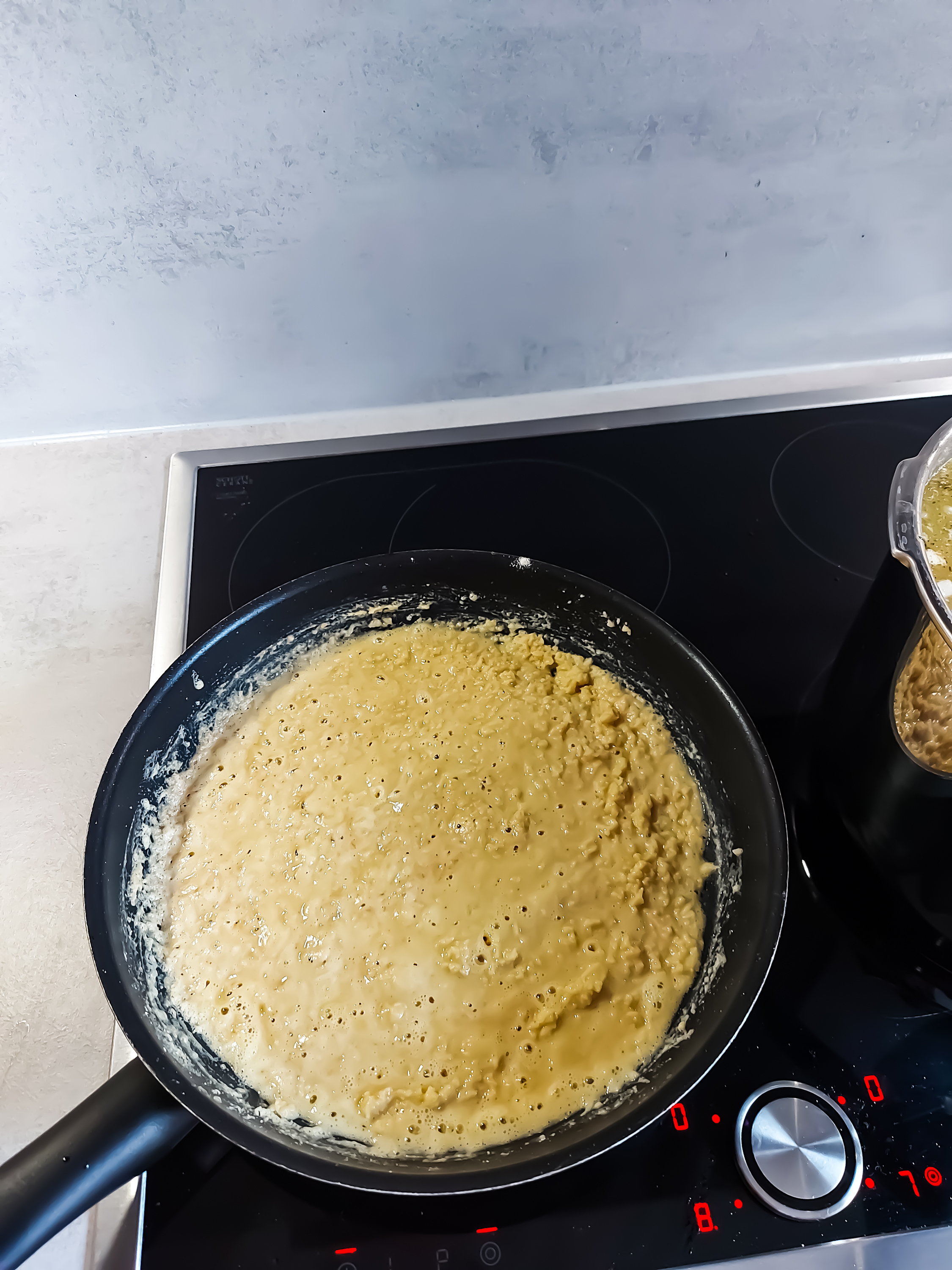 A frying pan on a stovetop with a semiliquid batter cooking, showing bubbles forming on the surface, indicating it's being cooked