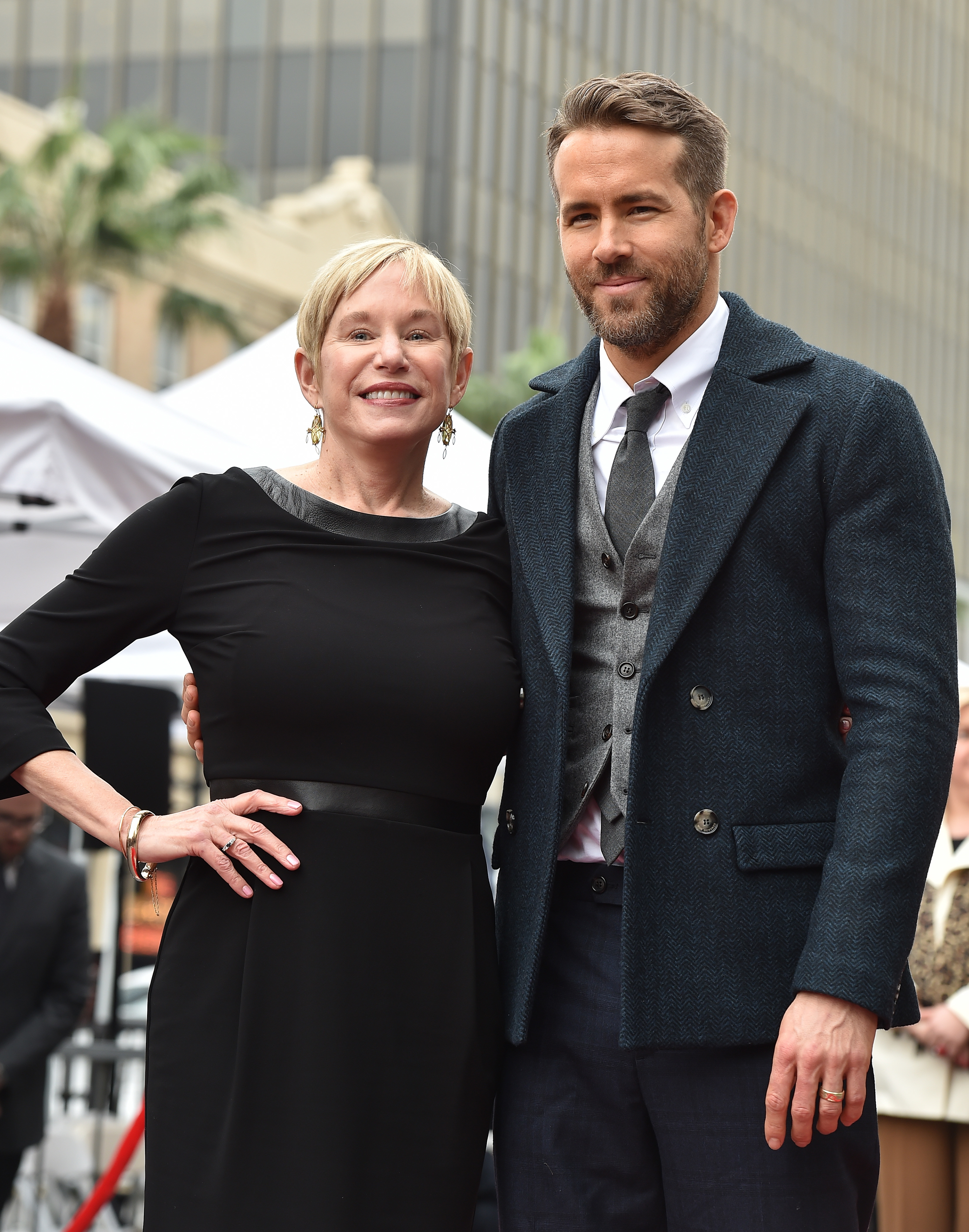 A woman in a black dress and Ryan Reynolds in a suit with an overcoat pose together outdoors, smiling