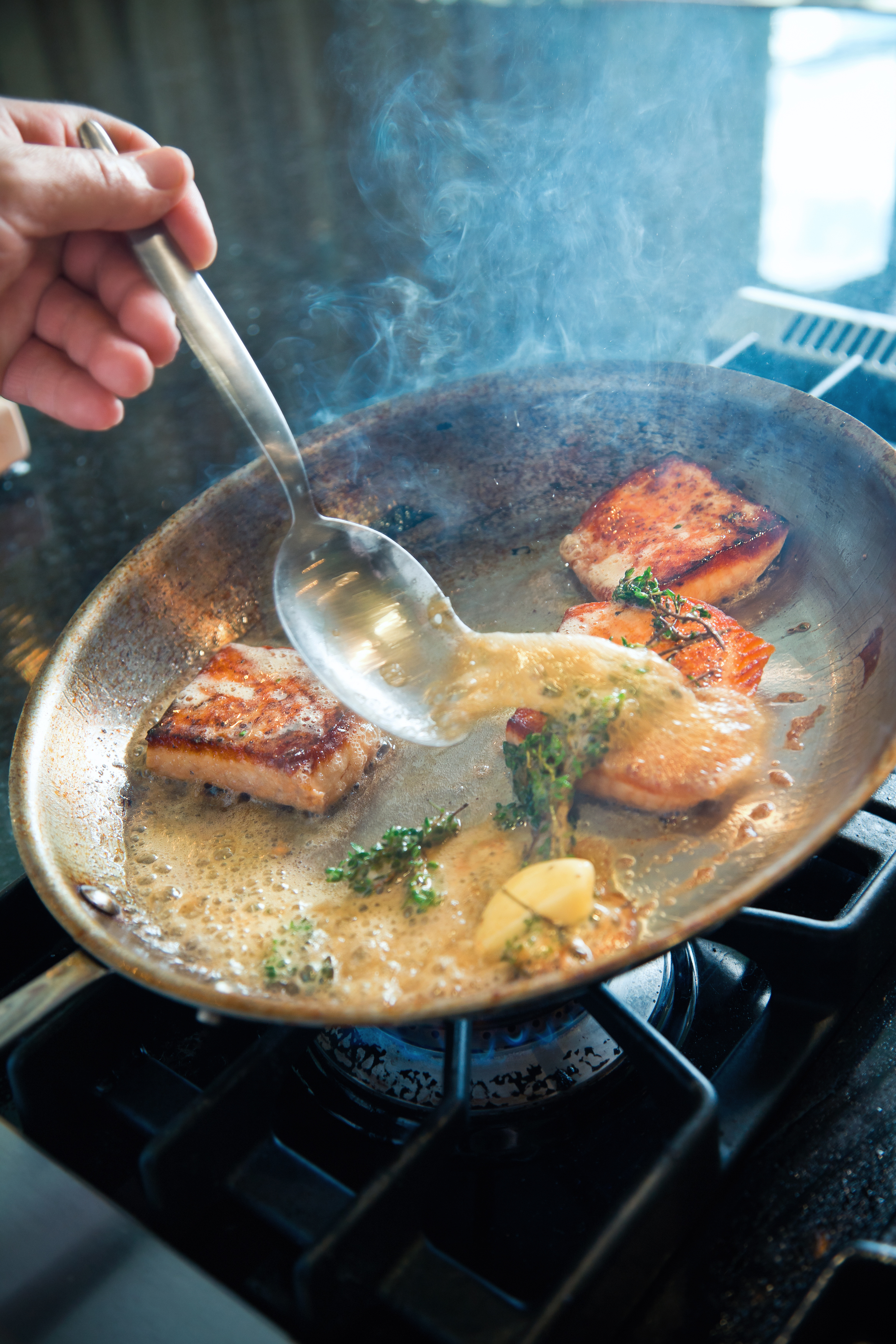 A hand uses a spoon to baste sauce over sizzling pieces of fish fillets in a frying pan on a stovetop