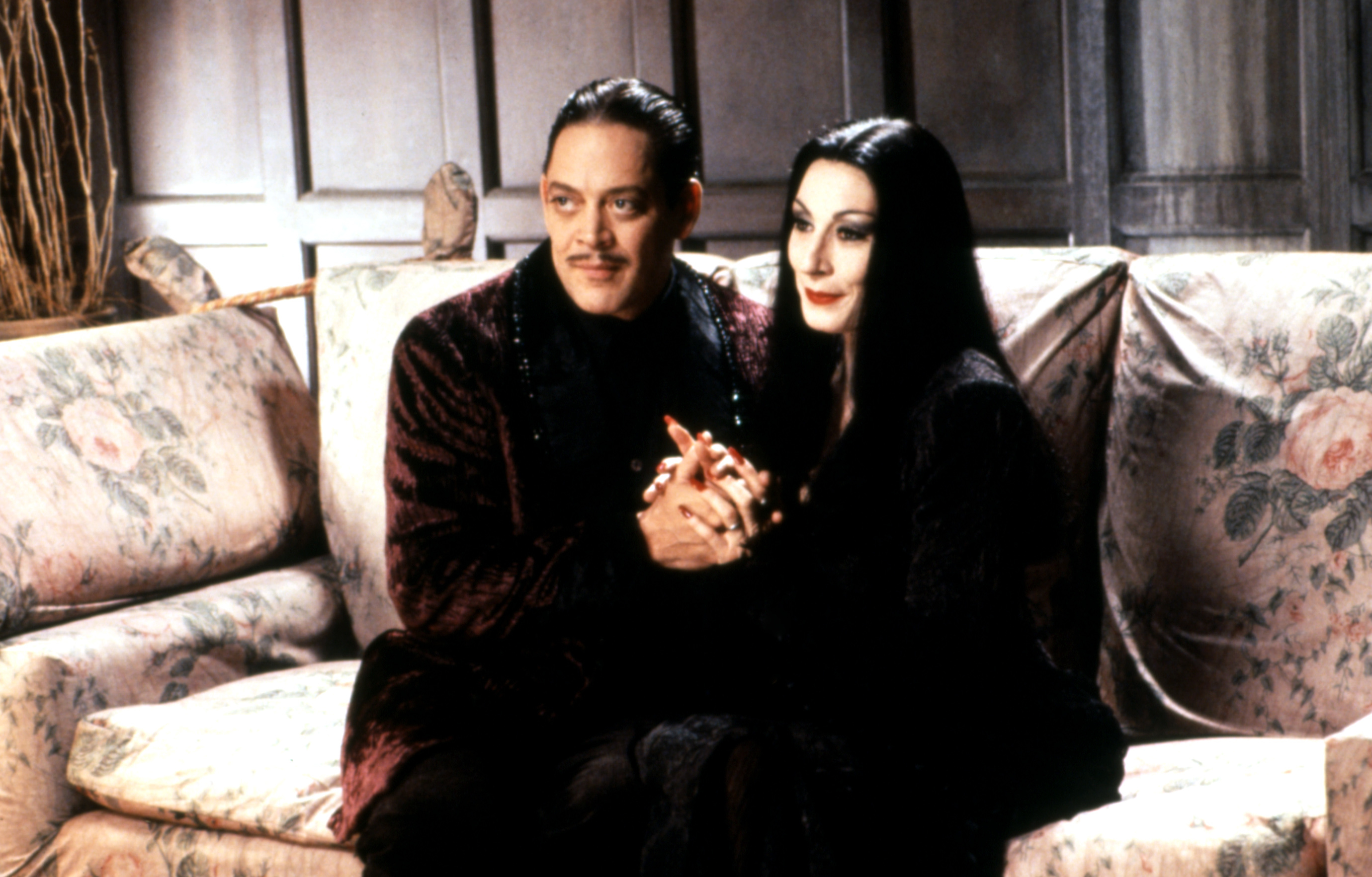 Gomez Addams and Morticia Addams, seated on a floral-patterned couch, holding hands and dressed in gothic-style attire