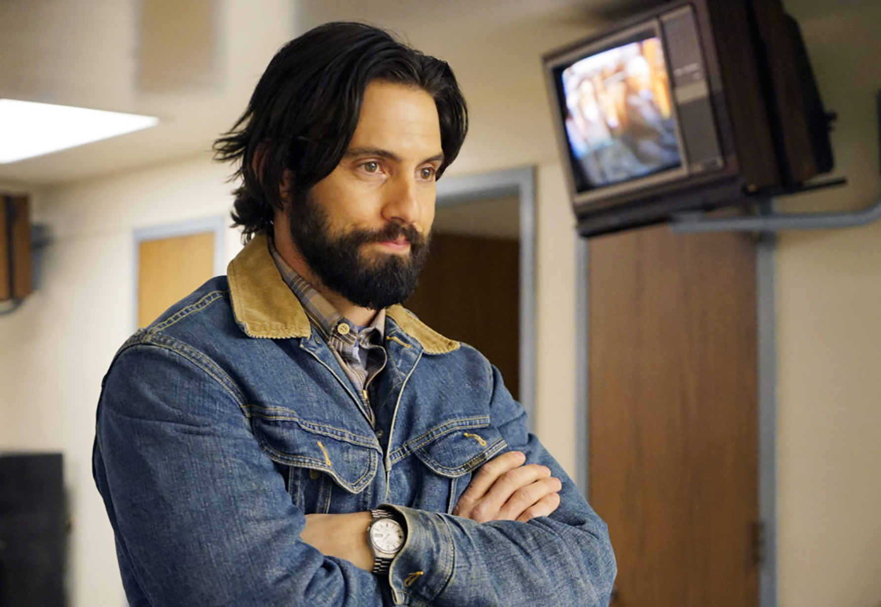 Milo Ventimiglia in a scene from “This Is Us,” wearing a denim jacket with a fur collar. A TV is visible in the background