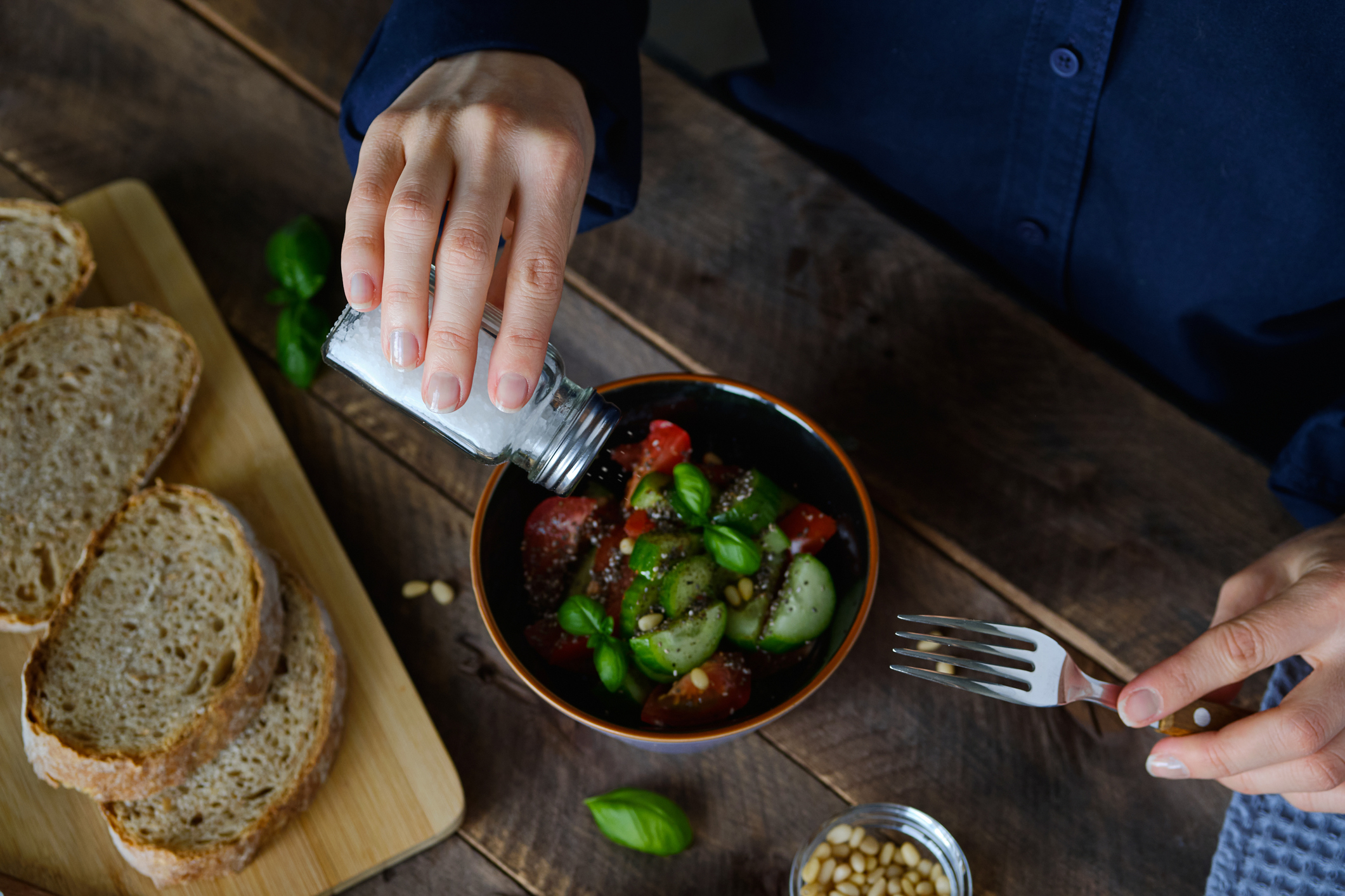 A person's hands sprinkling salt over a bowl of salad with sliced bread on the side on a wooden table