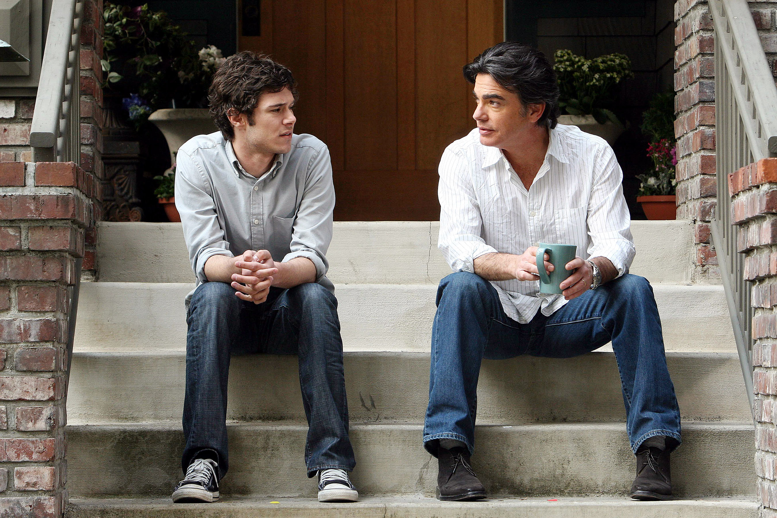 Adam Brody and Peter Gallagher sit on steps, talking. Adam wears a casual button-up shirt; Peter wears a similar shirt holding a mug