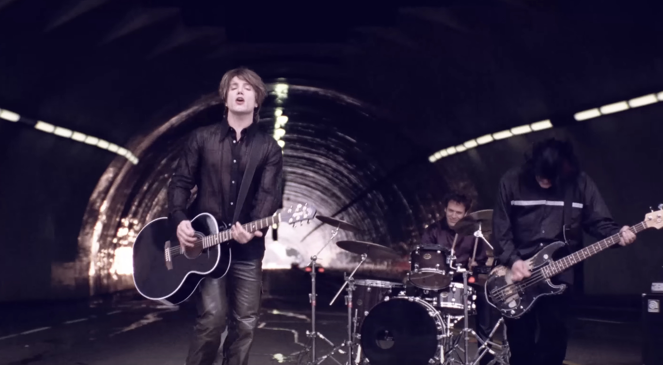 Lead singer of the Goo Goo Dolls plays a guitar, the drummer is at a drum set, and the bassist is playing the bass guitar