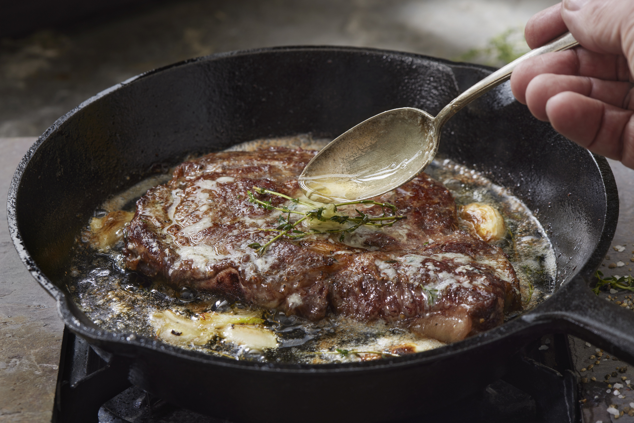 A close-up view of a steak being cooked in a cast-iron skillet, with a hand holding a spoon to baste it with melted butter and herbs