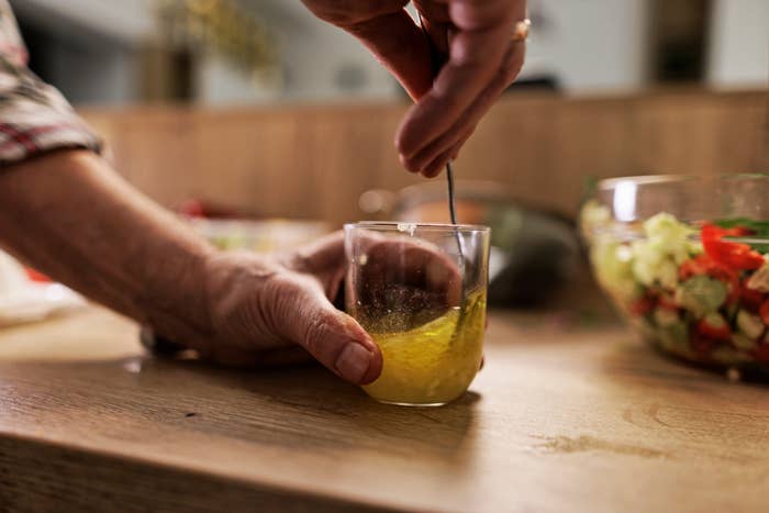 A pair of hands stirring a drink in a glass with a spoon on a wooden counter next to a bowl of chopped vegetables