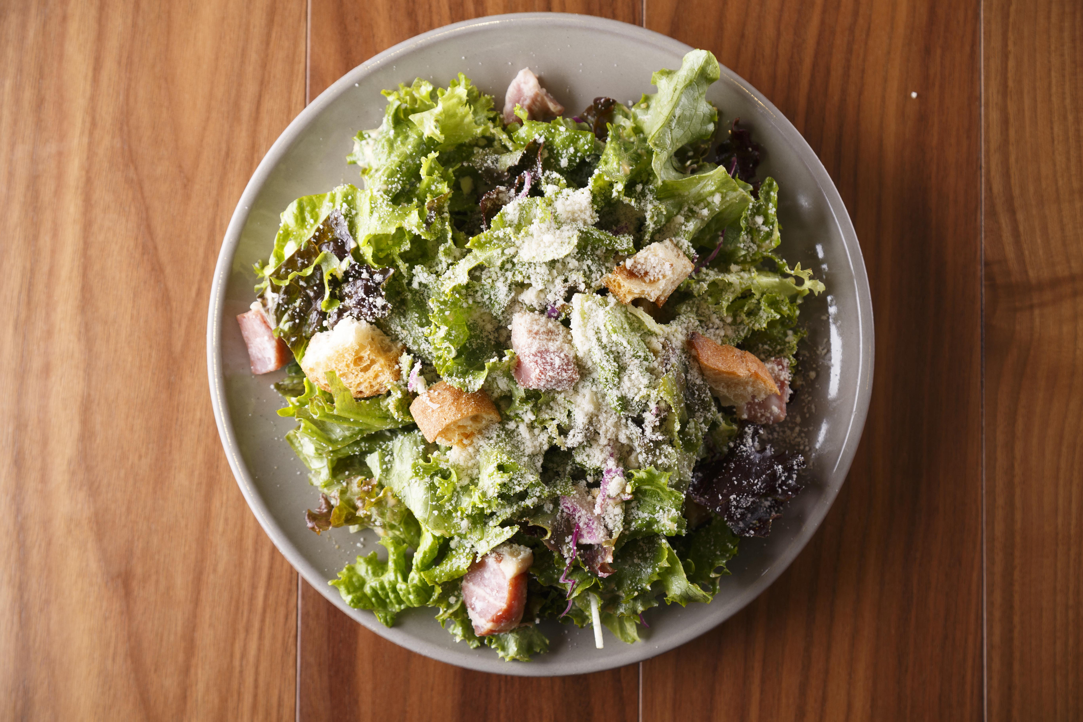 A fresh, mixed green salad with croutons and grated cheese on a plate, placed on a wooden table