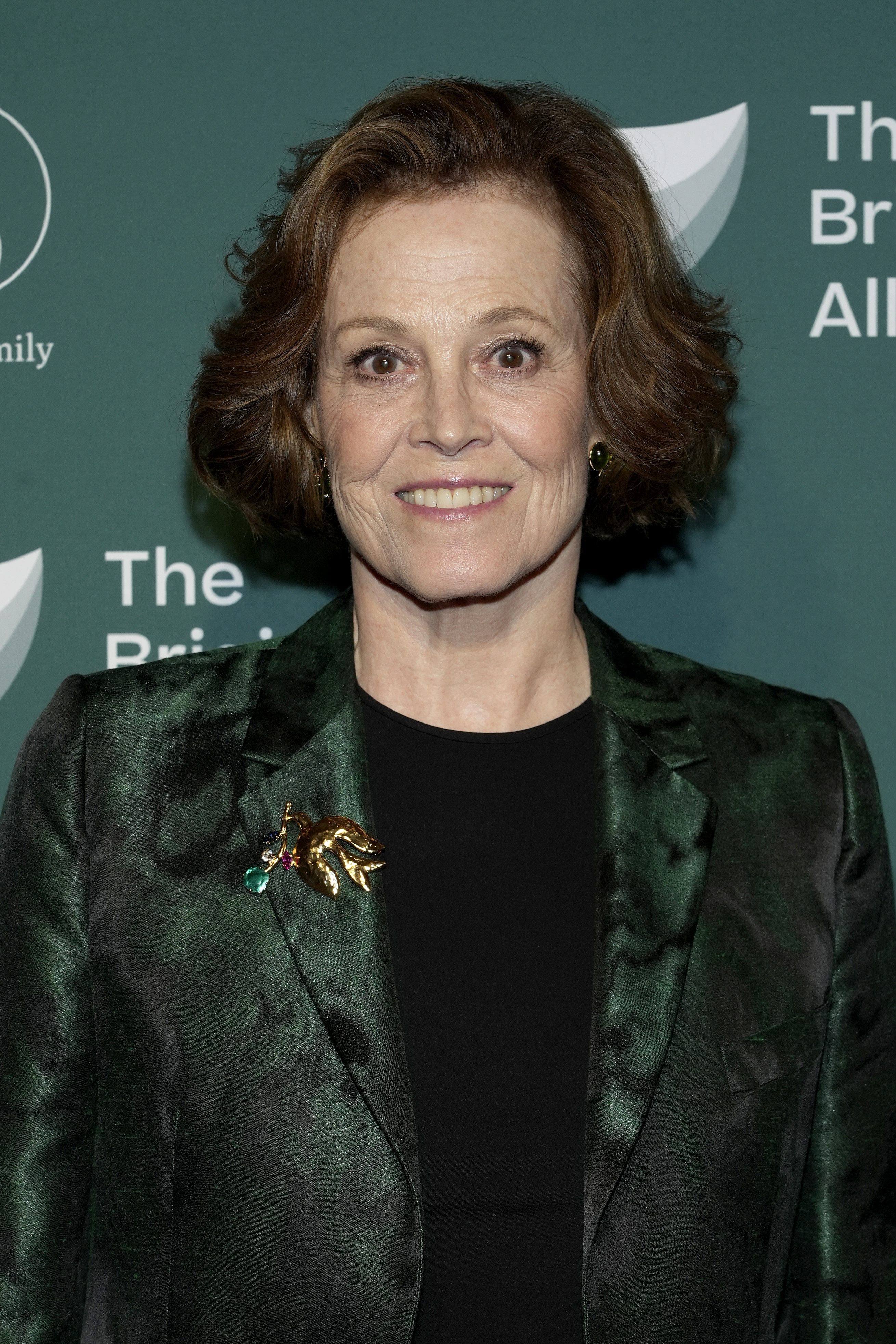 Sigourney Weaver wearing a dark blazer with a brooch, smiles at an event backdrop