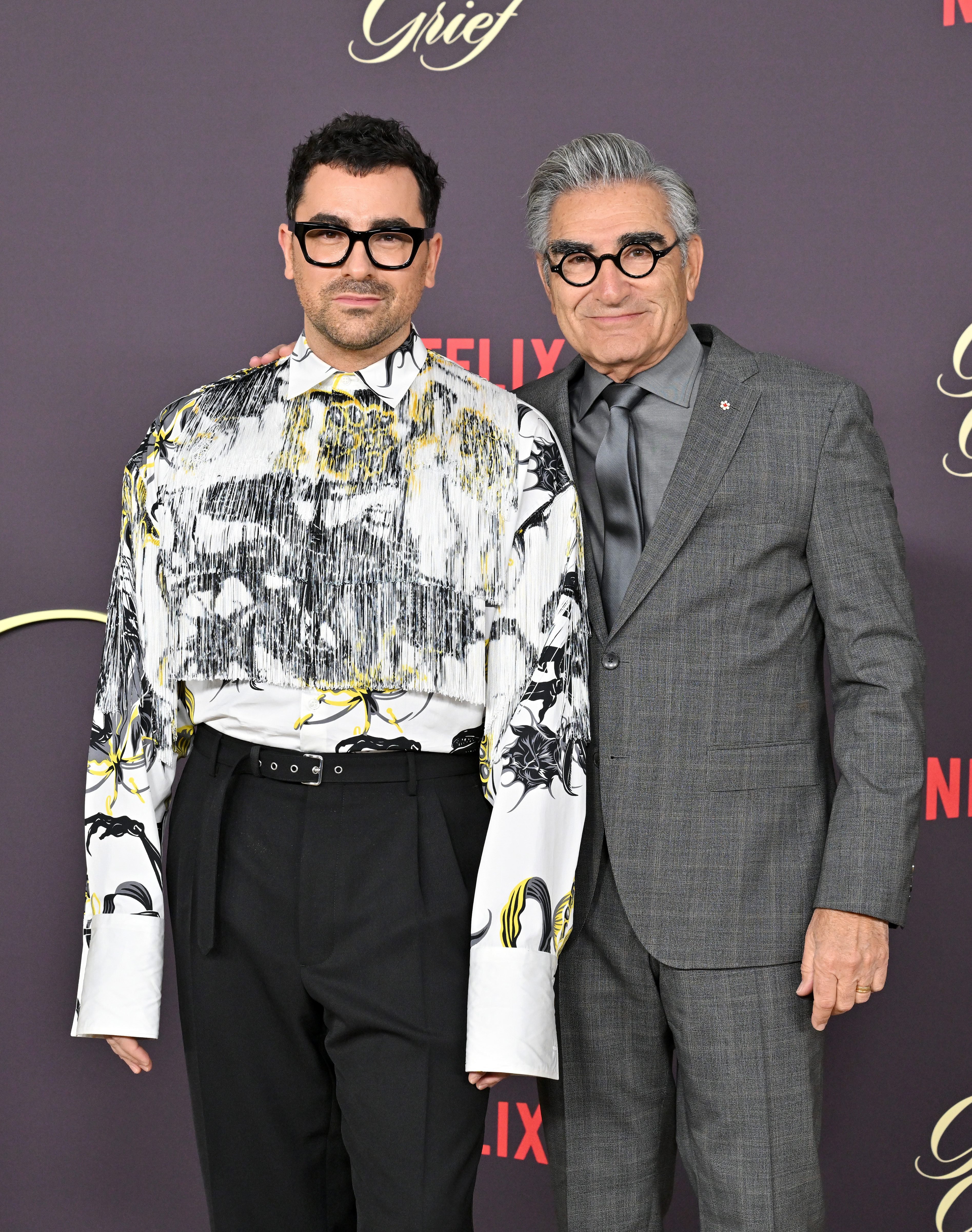 Dan Levy and Eugene Levy at a Netflix premiere, Dan wearing an abstract-patterned shirt and black pants, Eugene in a gray suit and tie