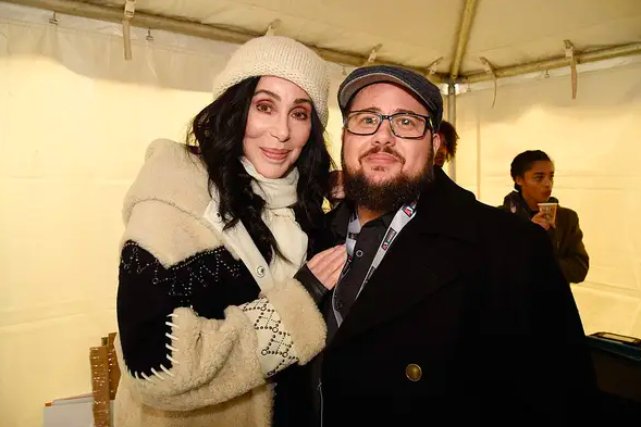 Cher and Chaz Bono smiling, standing together inside a tent at an event. Cher is wearing a cozy coat and a beanie, while Chaz is in a suit with a hat and glasses