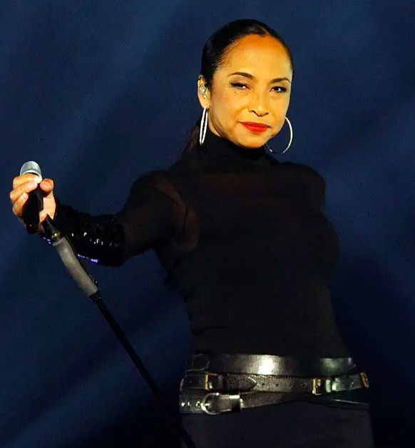 Sade Adu holds a microphone on stage, wearing a sleek black turtleneck and black pants with stylish belts, while performing