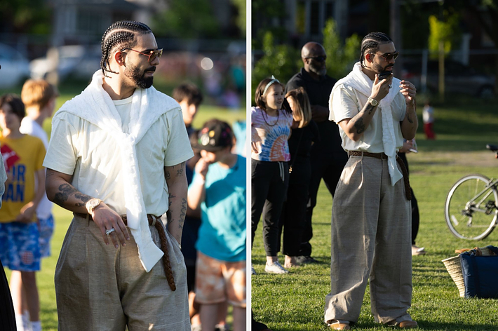 Man with braided hair and sunglasses wears a casual white shirt with a white sweater draped over his shoulders and loose, beige trousers at an outdoor event