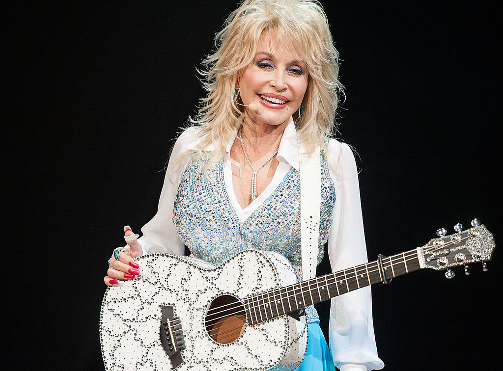 Dolly Parton, dressed in a sparkly vest and white shirt, smiles while holding a patterned acoustic guitar