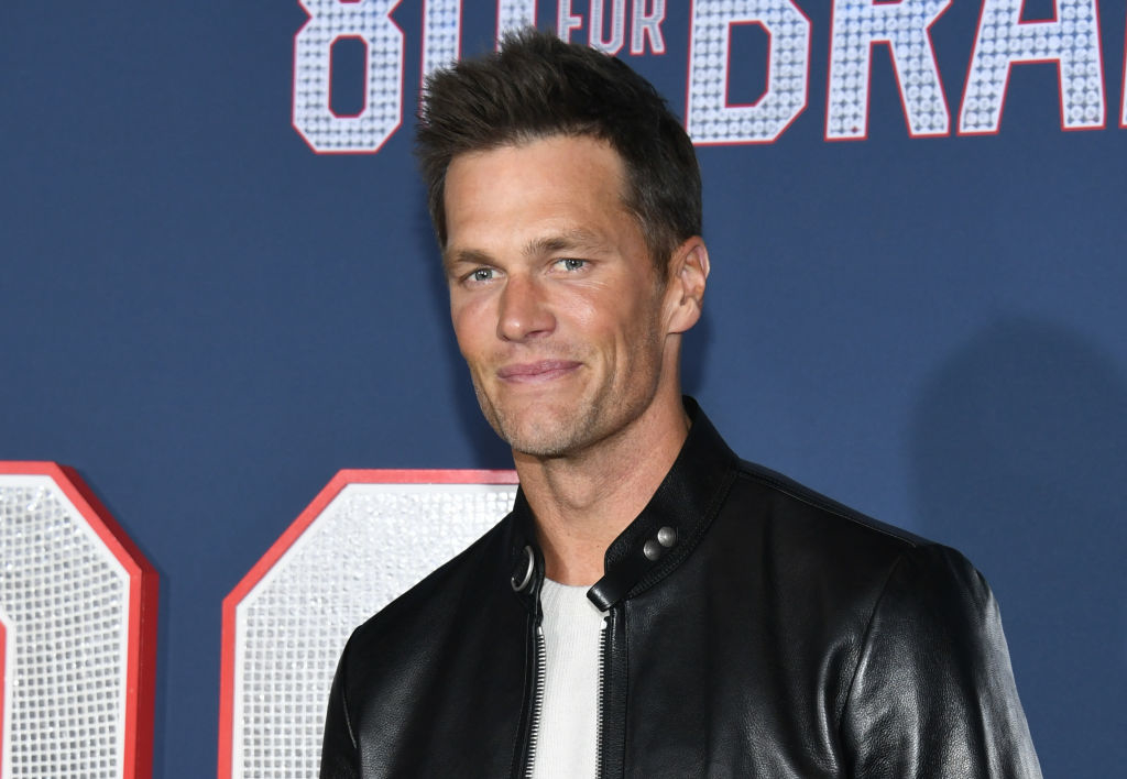 Tom Brady at an event, wearing a black leather jacket over a white top, smiling. The backdrop has the text "80 for Brady."