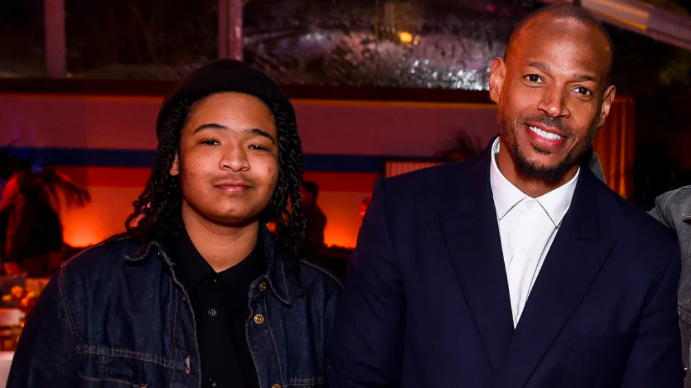 Marlon Wayans and his child pose together at an event; Marlon is wearing a navy suit and white shirt, while his child wears a denim jacket and black shirt
