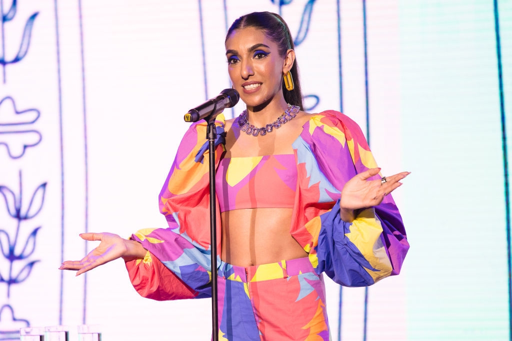 Rupi Kaur speaks at a podium, dressed in a colorful, geometric-patterned crop top and puffed sleeves outfit, with hair pulled back