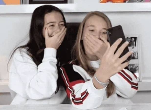 Two girls in casual clothing laugh and cover their mouths while taking a selfie. Shelves with various photos and awards are visible in the background