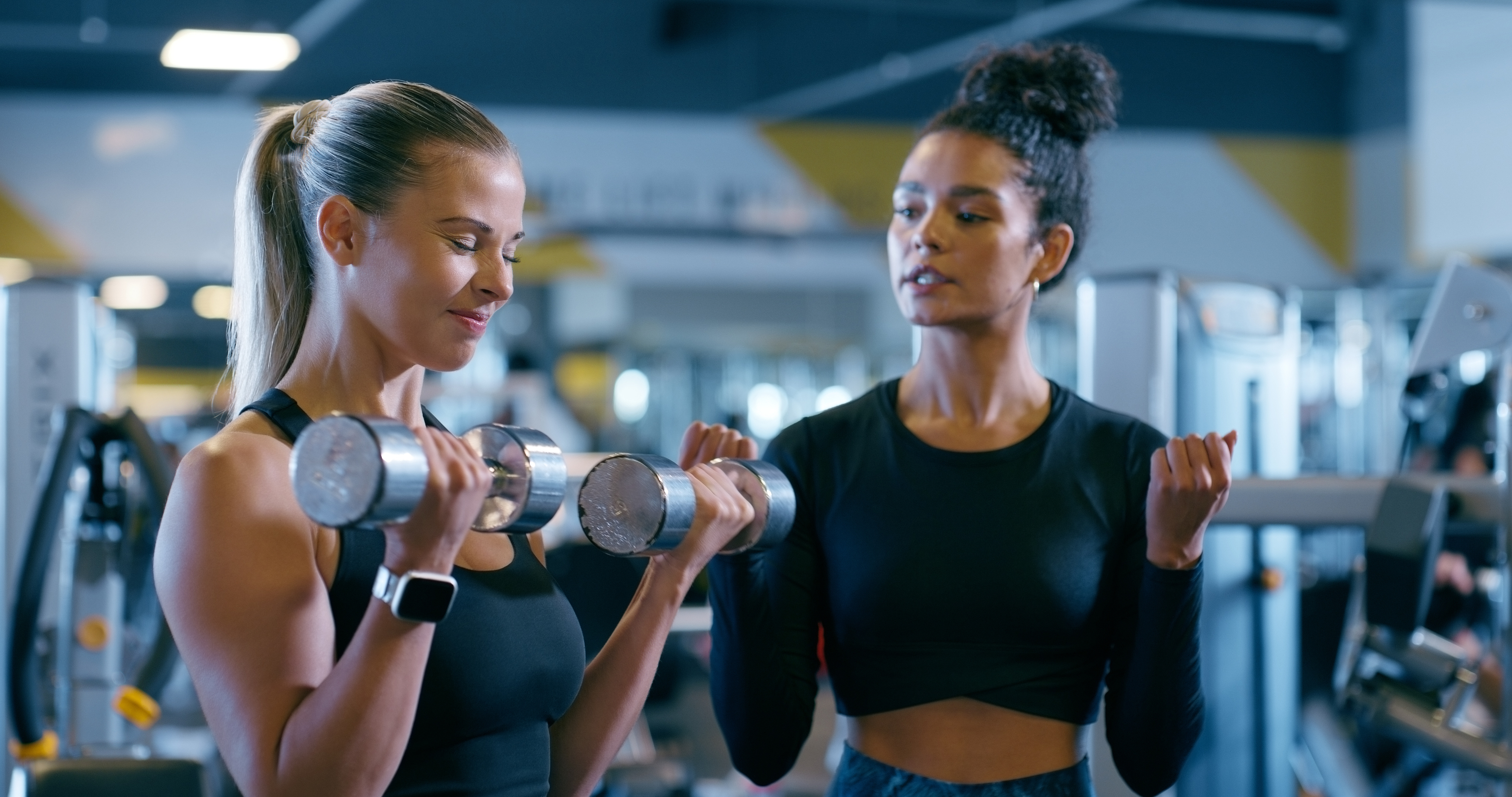 Two women in a gym, one lifting dumbbells while the other, possibly a trainer, observes. Both wear athletic clothing