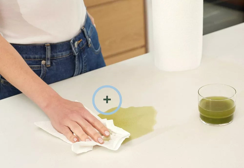 A person cleans a green liquid spill on a countertop using a paper towel next to the text: "50% Thicker & More Absorbent"