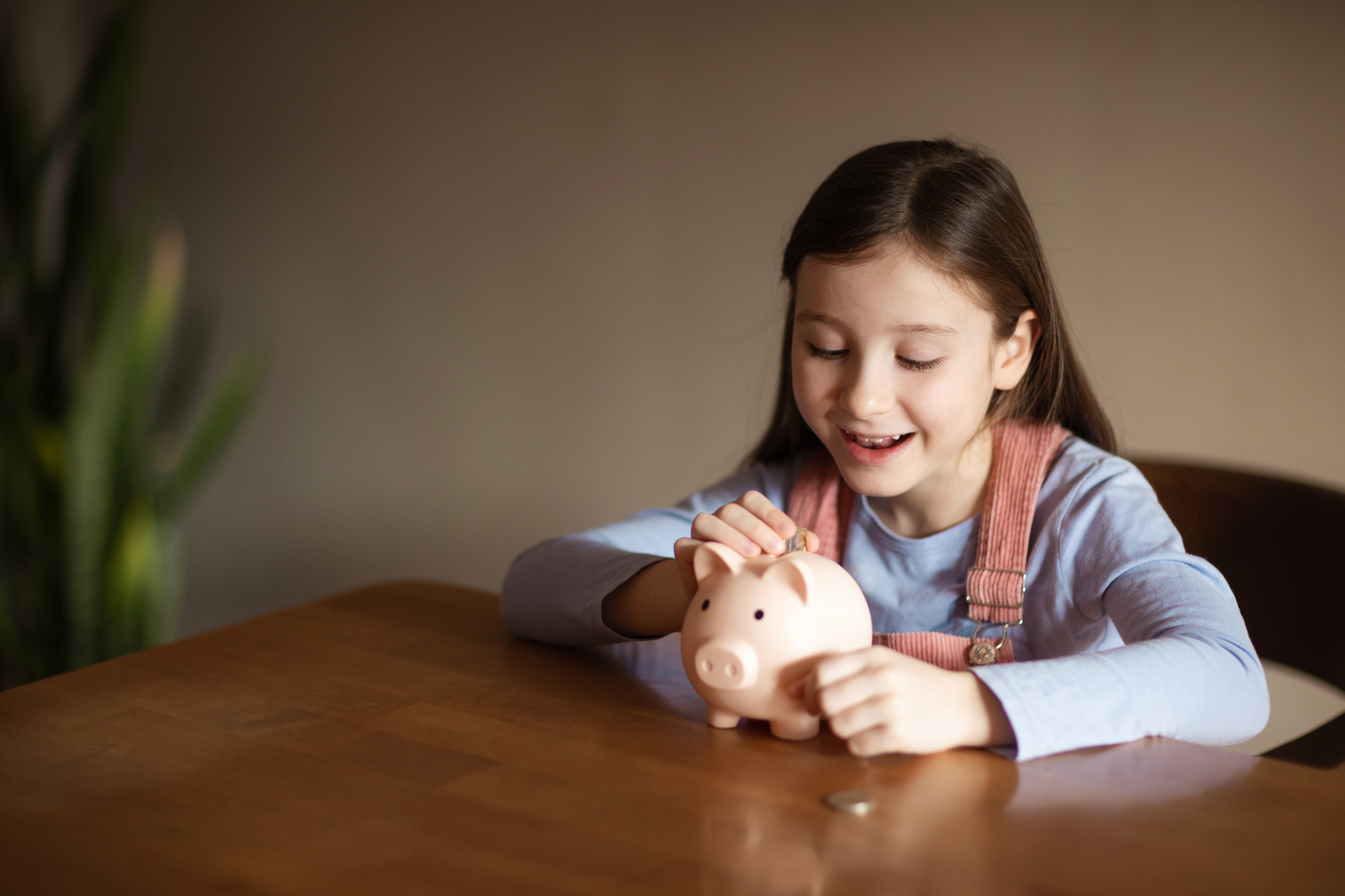 A young girl smiles while placing a coin into a piggy bank at a wooden table