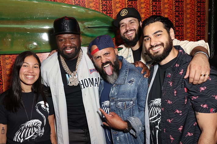 50 Cent and four other smiling people, three men and one woman, pose together at a lively event. Casual clothing is mixed with a baseball jersey