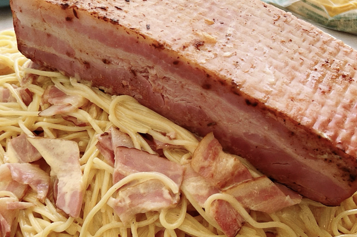 スパゲッティの上に大きなベーコンのブロックが乗っている料理の画像。