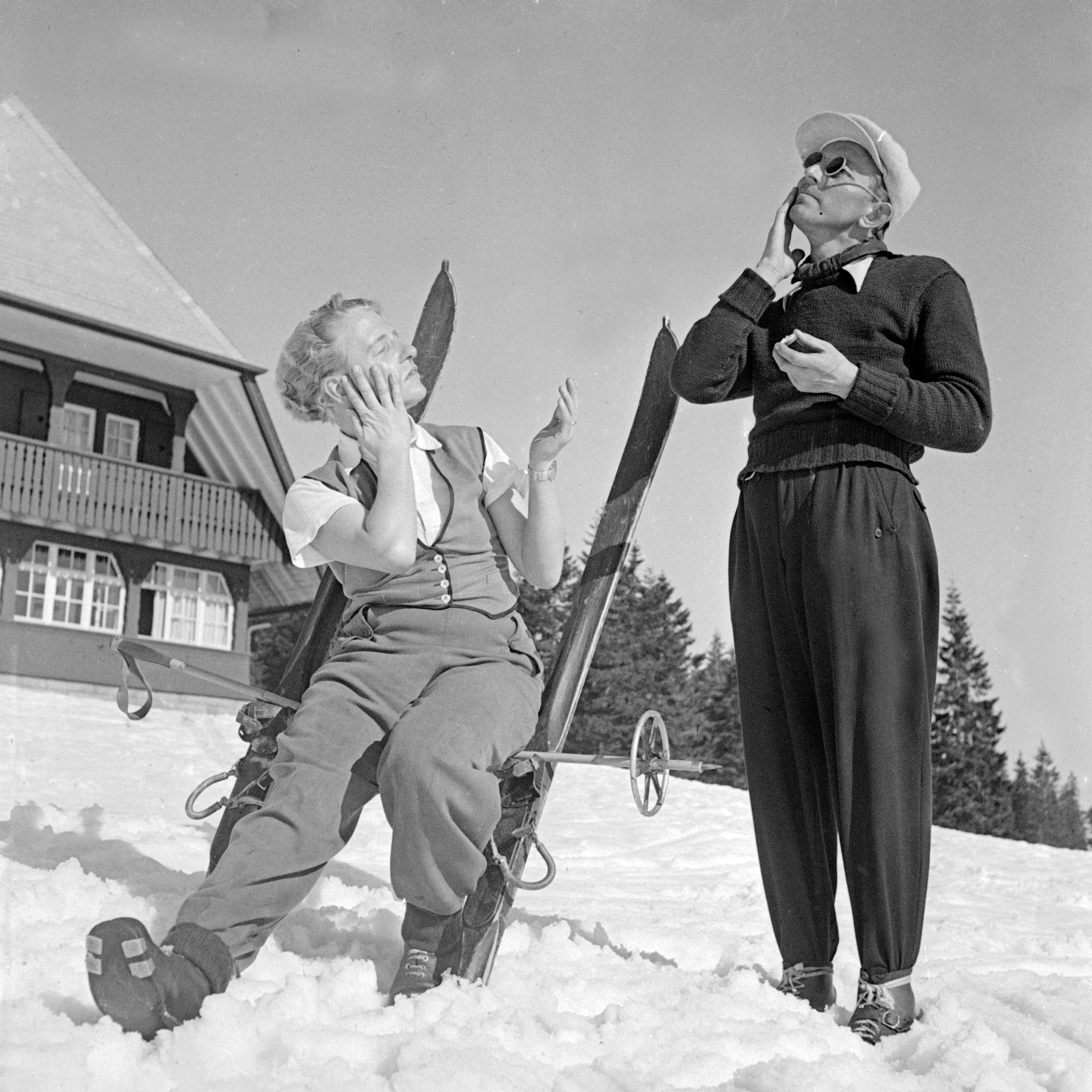 Two people dressed in vintage ski attire, one seated with skis and the other standing, in a snowy outdoor setting by a cabin