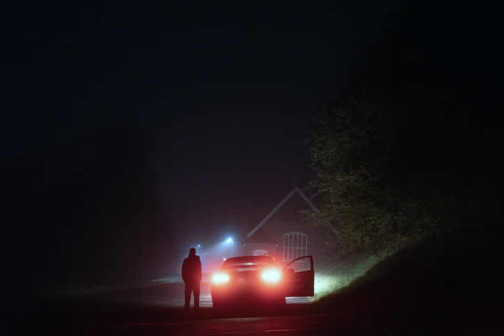 Silhouette of a person standing near a car with headlights on in a dark, foggy area beside a small building