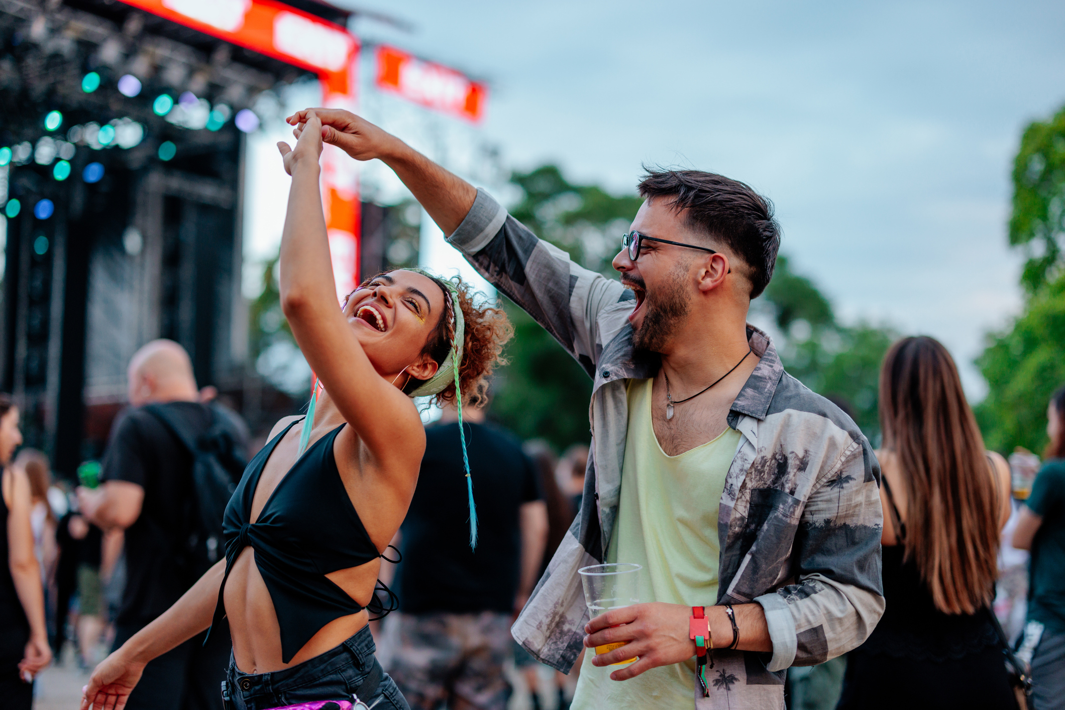 Couple dancing joyfully at an outdoor festival, woman in a stylish crop top, man in a casual shirt with a drink in hand, with a stage in the background