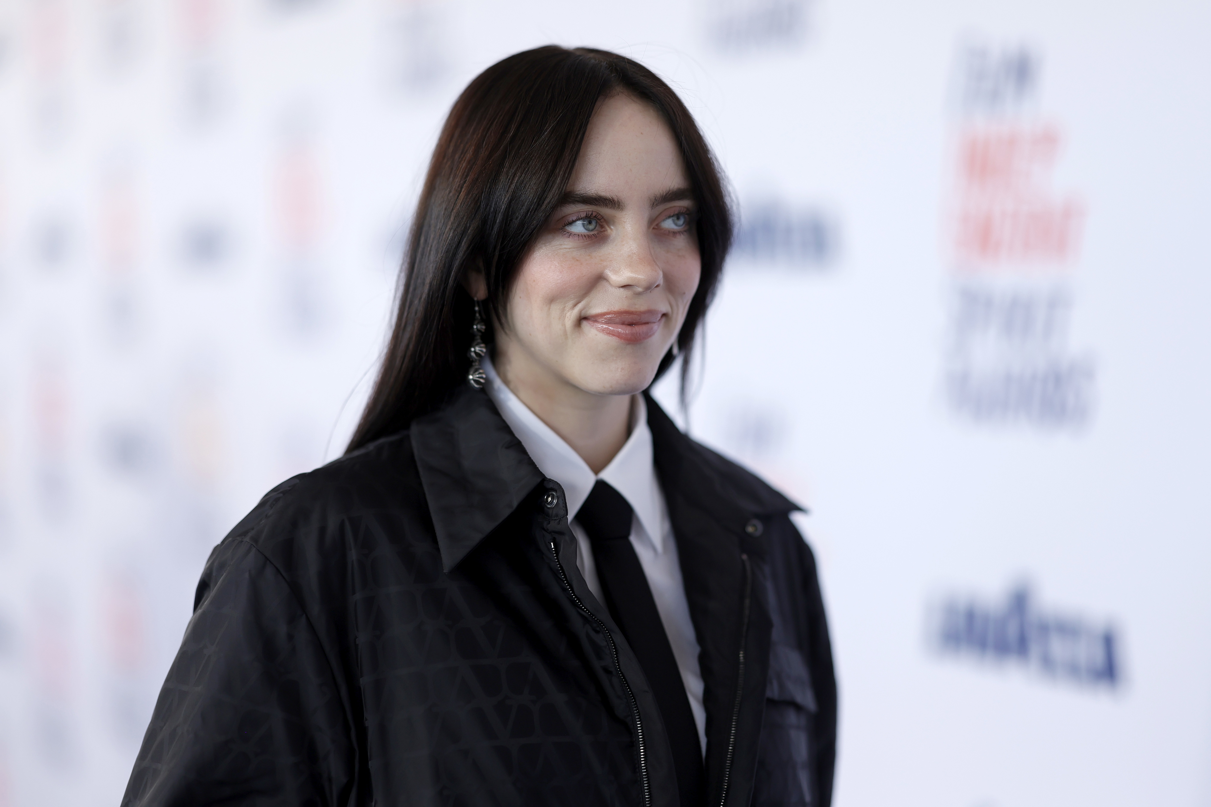 Billie Eilish on the red carpet, wearing a black jacket and white shirt with a black tie, smiling slightly