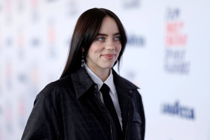 Billie Eilish on the red carpet, wearing a black jacket and white shirt with a black tie, smiling slightly