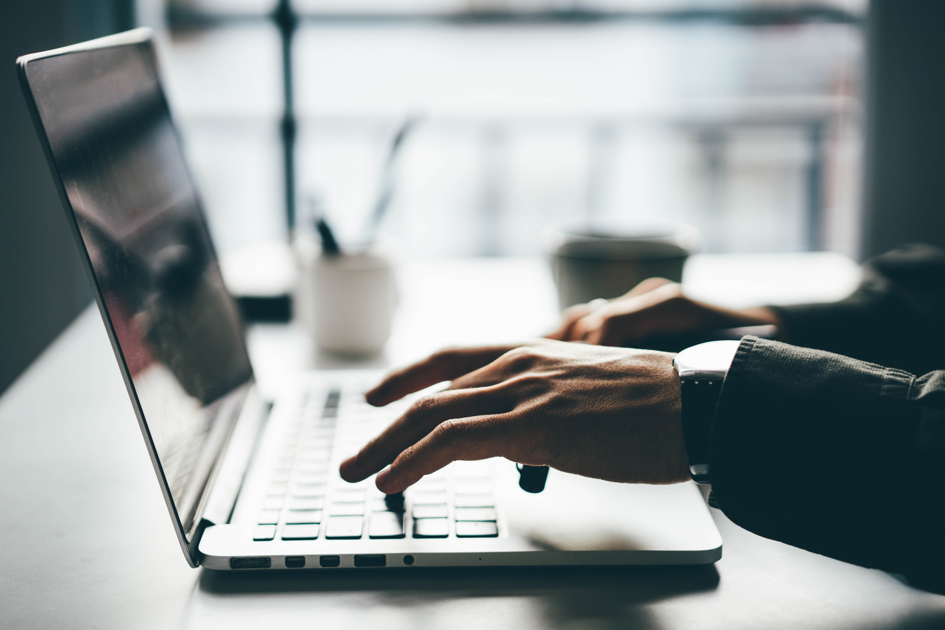 Close-up of hands typing on a laptop, with a cup and blurry background