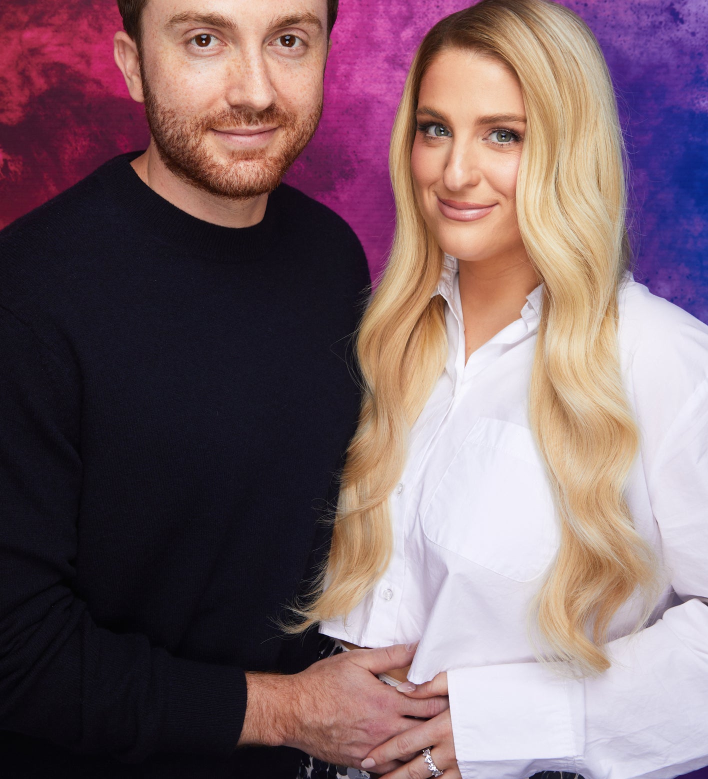 Meghan Trainor and Daryl Sabara pose together, smiling. Daryl wears a black sweater. Meghan wears a white blouse and a sequined skirt, her hand on her baby bump