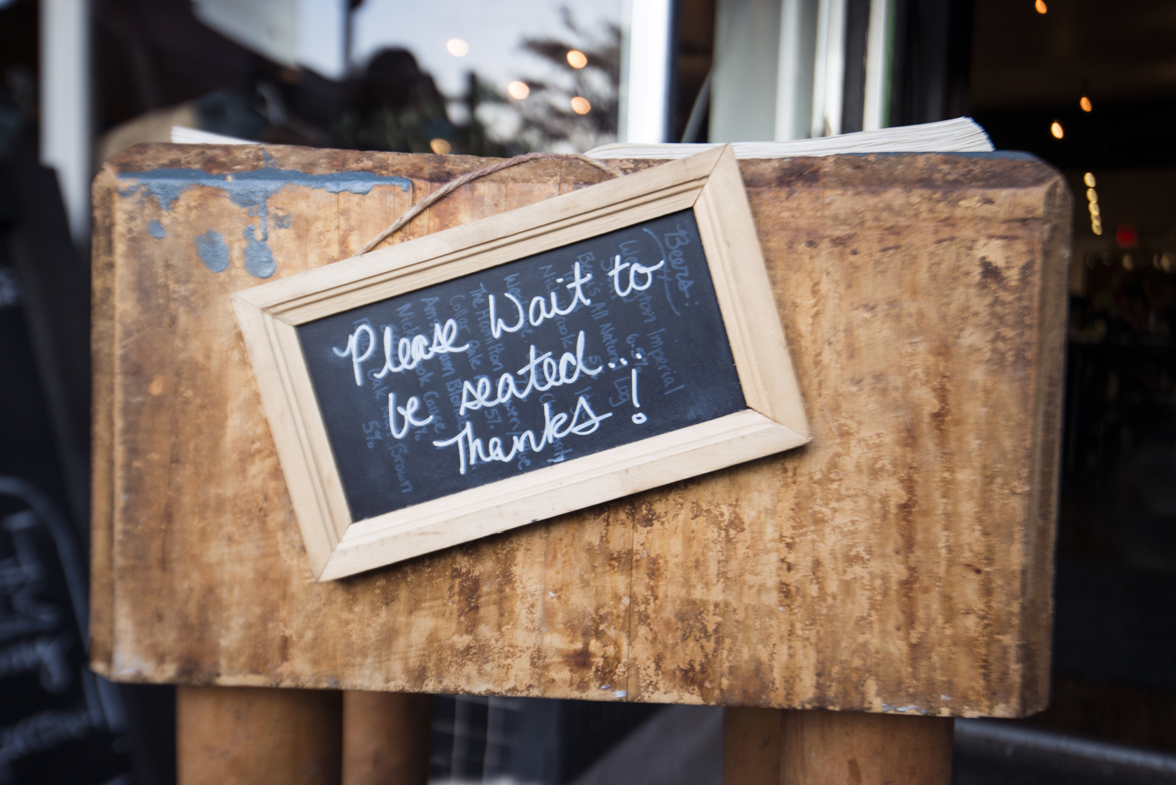 Chalkboard sign on a wooden stand reads, "Please wait to be seated. Thanks!" Image likely taken at a restaurant entrance