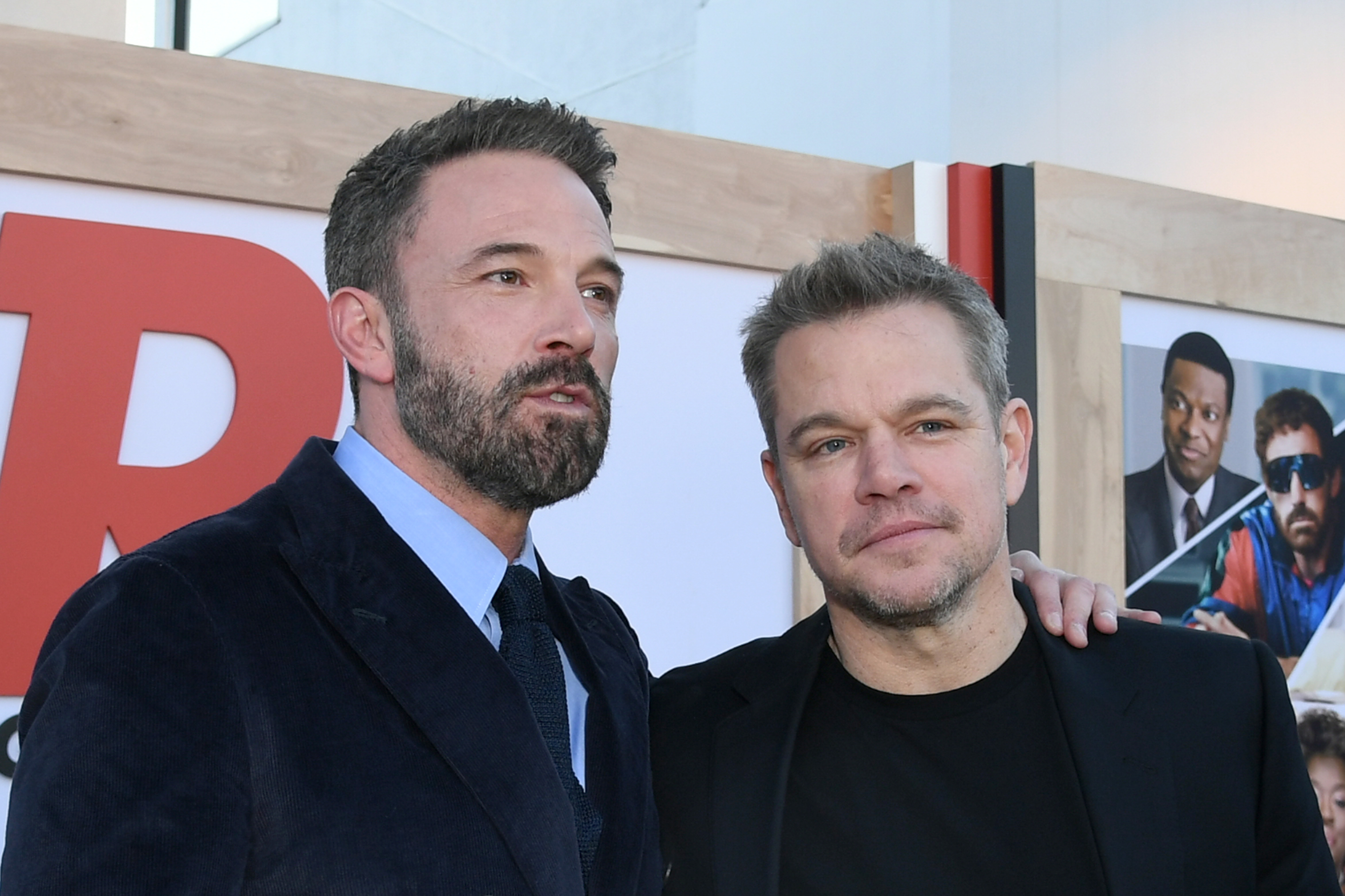 Ben Affleck in a suit and tie, and Matt Damon in a black shirt and blazer, pose together at an event