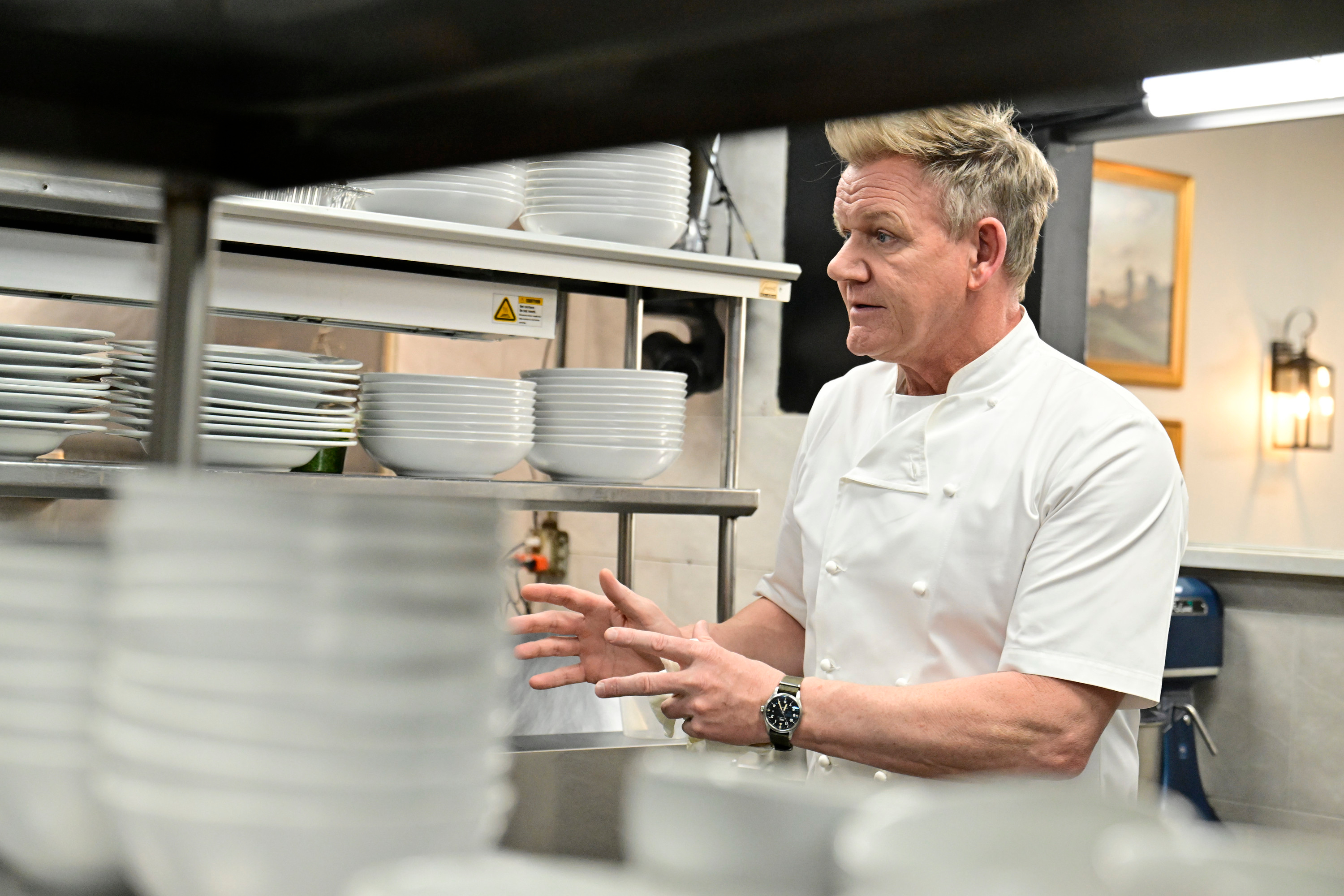 Gordon Ramsay stands in a kitchen, dressed in a white chef's jacket, actively gesturing with his hands near shelves stacked with plates