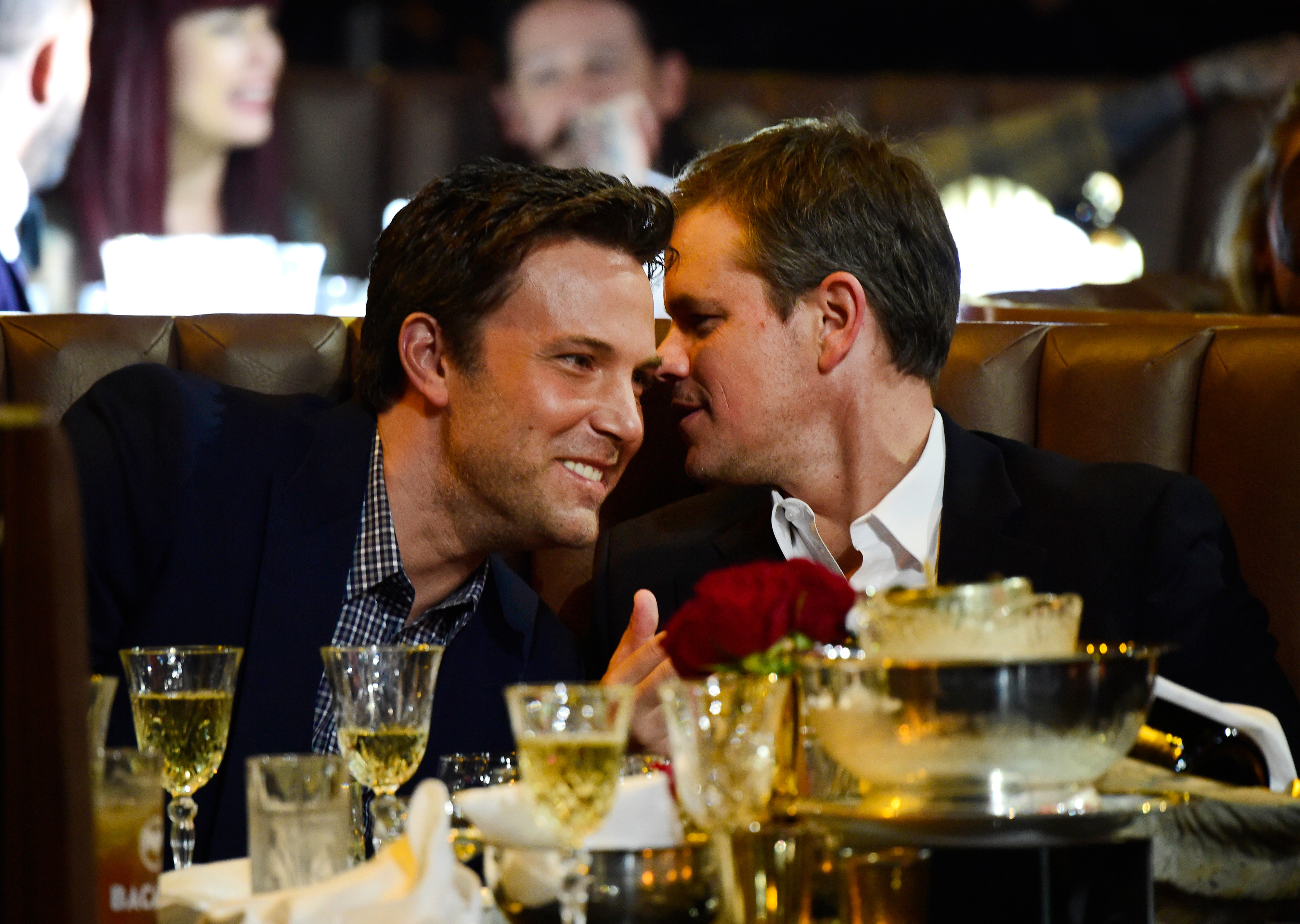 Ben Affleck and Matt Damon are sitting close together at a formal event, smiling and deep in conversation, surrounded by glasses and wine