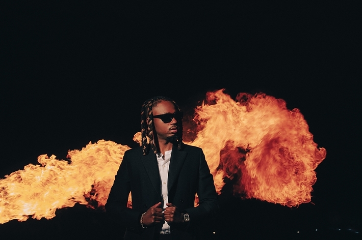 Musician in a dark suit and sunglasses stands confidently in front of large flames, creating a dramatic backdrop