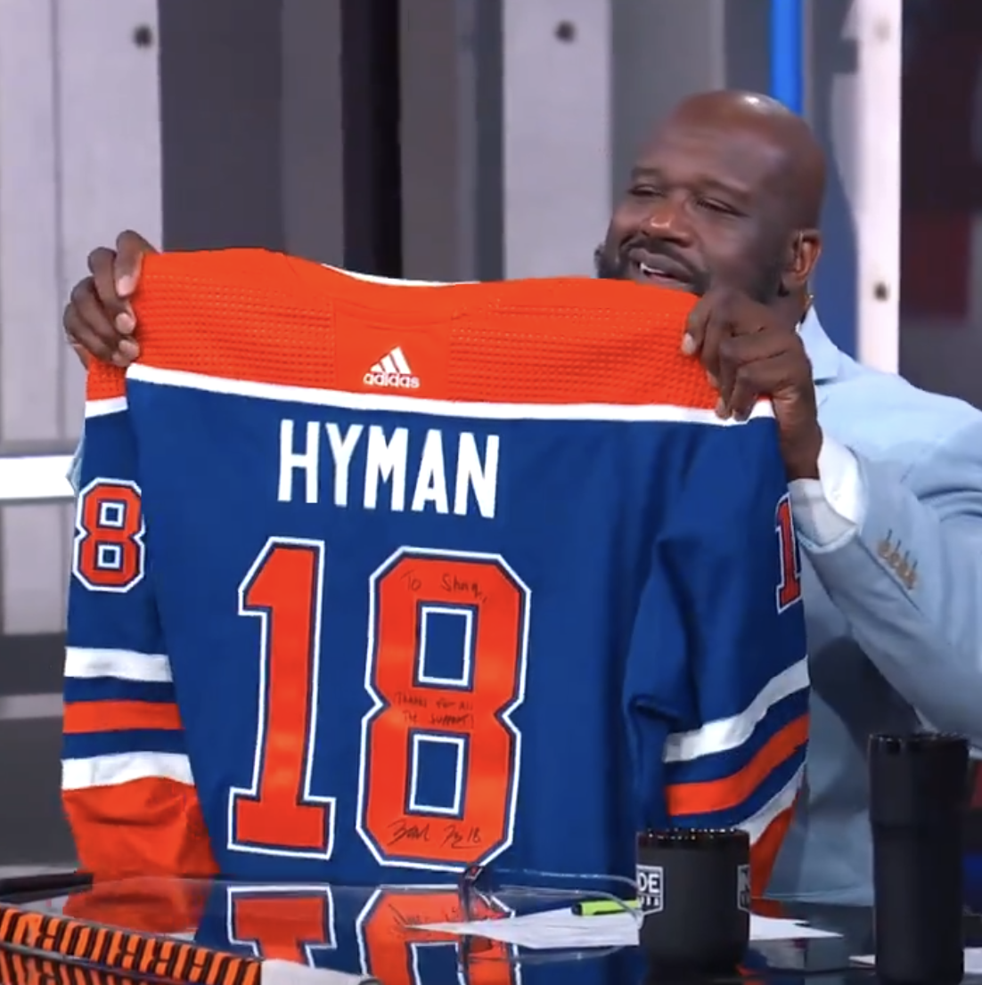 Shaquille O'Neal holds up a hockey jersey with the name &quot;Hyman&quot; and the number 18, which includes a handwritten message