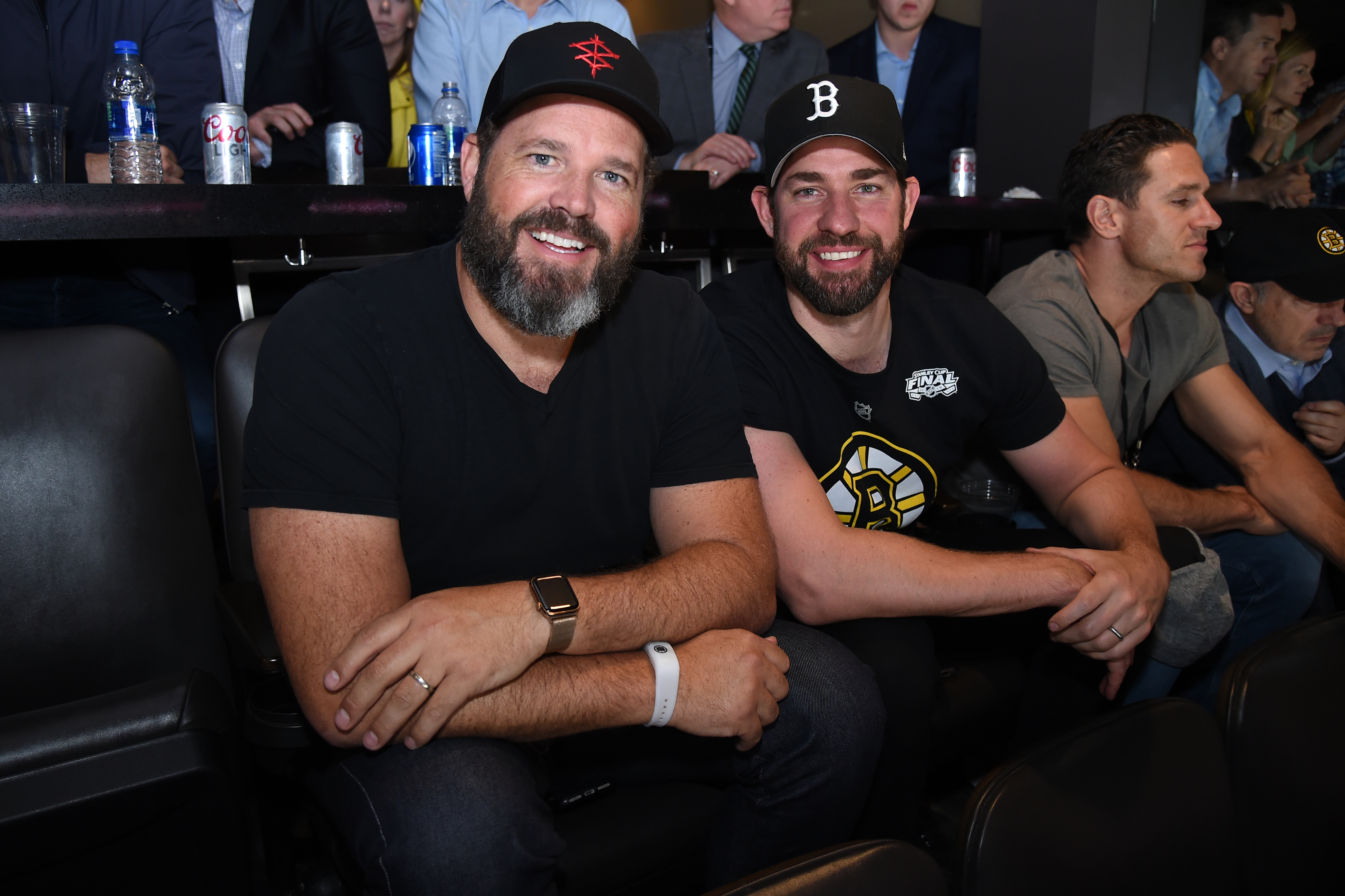 Two men, wearing casual black attire and baseball caps, smile and sit together at a sports event. The man on the right wears a Boston Bruins shirt
